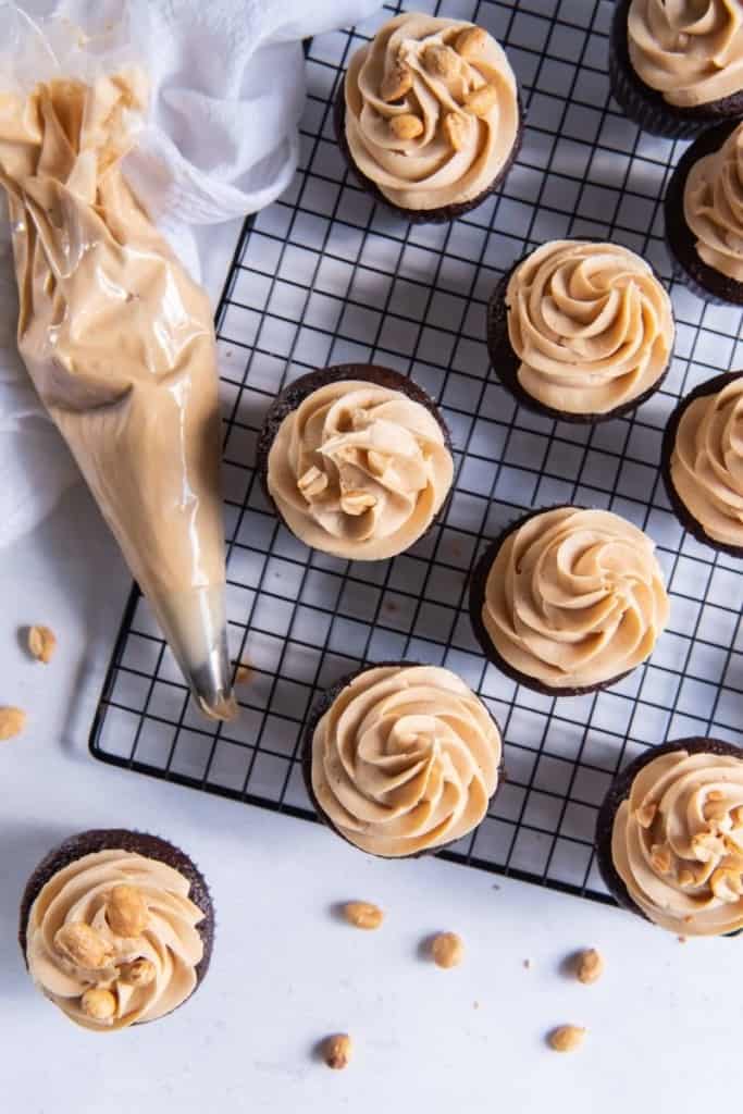 Peanut butter frosting on top of chocolate cupcakes on a cooling rack and a piping bag next to it