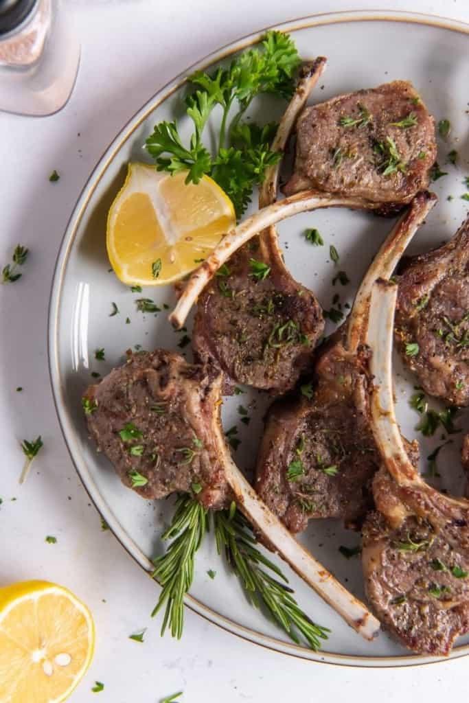 Lamb chops on a white plate with green parsley and rosemary garnish and lemon wedges