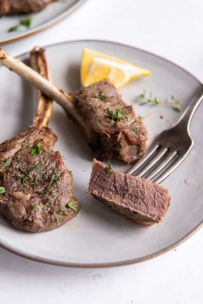 Lamb chops on plate with lemon wedge and one lamb chop piece cut onto a fork