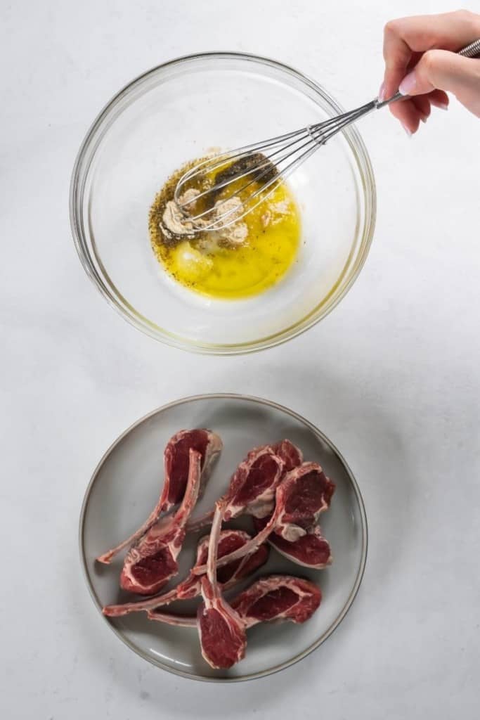 Marinade ingredients being mixed together in a bowl with a whisk and lamb chops on a plate below