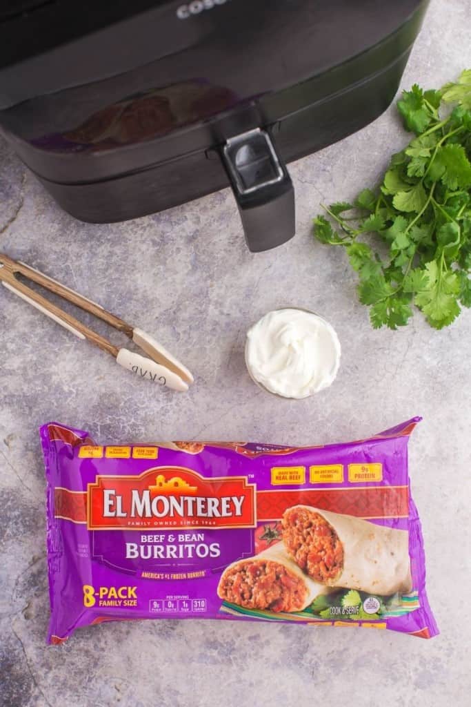 El Monterey burritos in front of an air fryer with tongs, sour cream, and cilantro