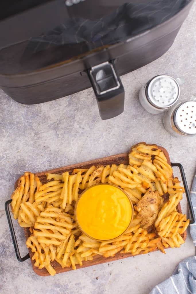 Overhead photo of cooked frozen waffle fries on a serving tray with nacho cheese in front of an air fryer