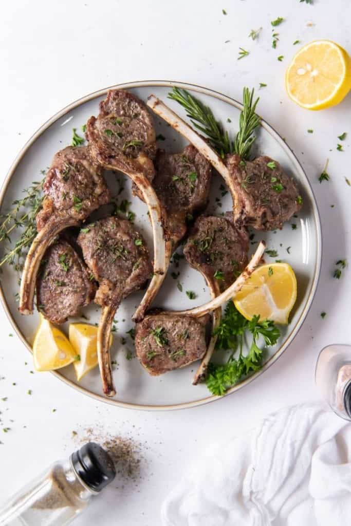 Overview shot of lamb chops served together on a white plate with lemon wedges and green garnishes