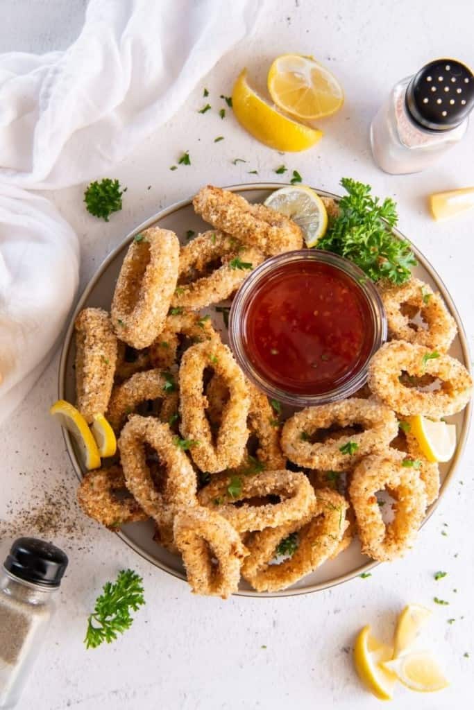Zoomed out view of the breaded calamari on a serving platter with sweet chili sauce and lemon wedges and green garnish around it