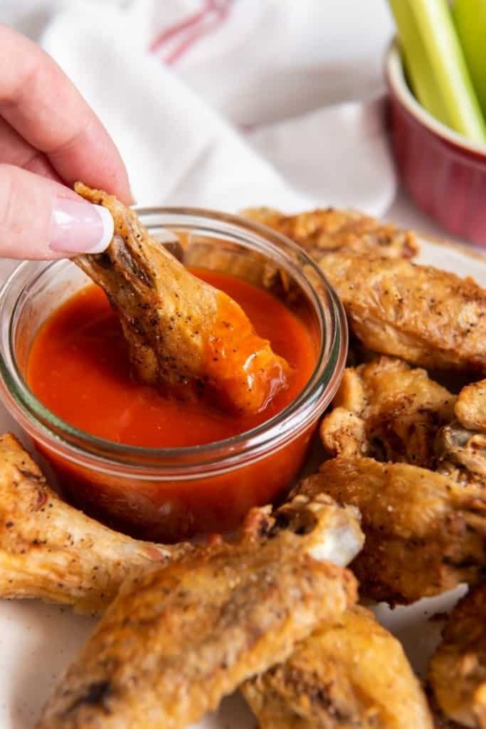 Chicken wing being dipped into buffalo sauce with other wings on plate