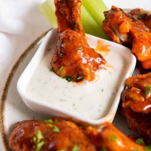 Air Fryer Frozen Chicken Wings coated with buffalo sauce on a white plate with one drumstick being dipped into blue cheese