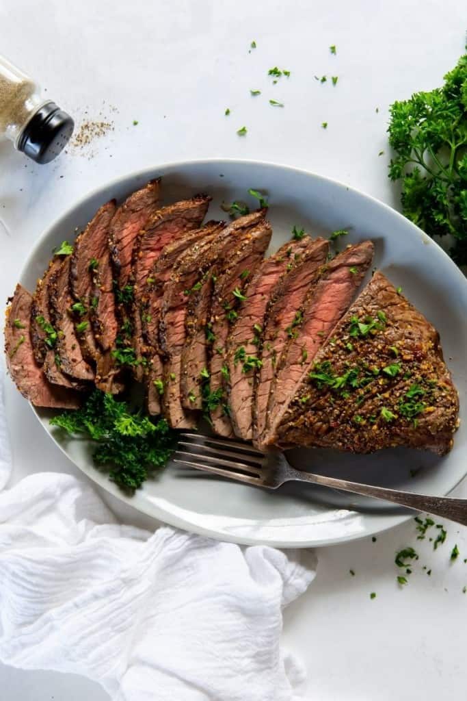 London broil on a white serving plate sliced with a fork and parsley on top