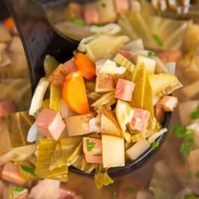 A ladle scooping out the soup with a lot of ham, cabbage, and potatoes on it.