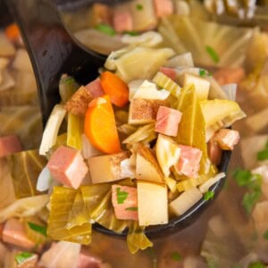 A ladle scooping out the soup with a lot of ham, cabbage, and potatoes on it.