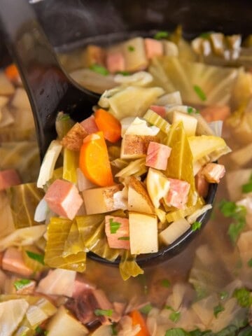 A ladle scooping out the soup with a lot of ham, cabbage, and potatoes on it.