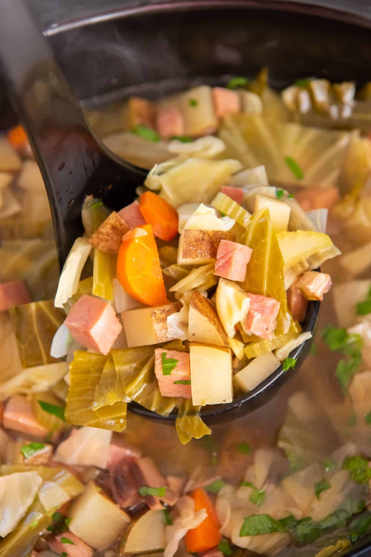 A ladle scooping out the soup with a lot of ham, cabbage, and potatoes on it.