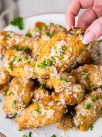 Hand reaching for one of many garlic parmesan wings on plate