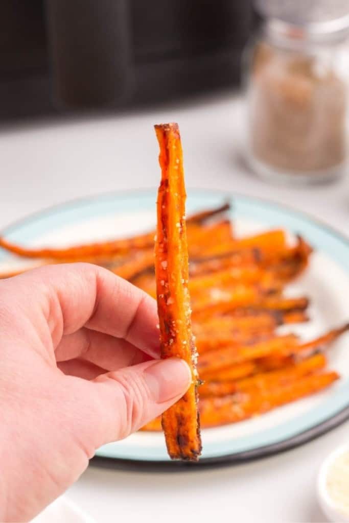 hand holding up a single carrot fry in front of plate of fries