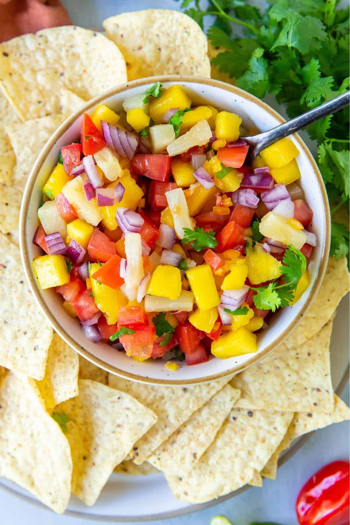 Mango pineapple salsa in a bowl with a spoon in it to scoop out some and surrounded by tortilla chips.