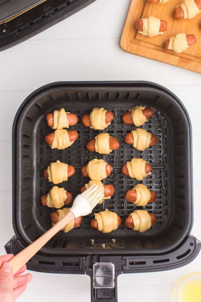 Pigs in a blanket in an air fryer basket about to be air fried and being basted with butter on top.