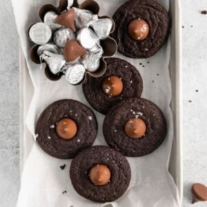 Overhead view of chocolate kiss cookies on parchment paper in a storage tin and a little bowl of Hershey kisses on the side.