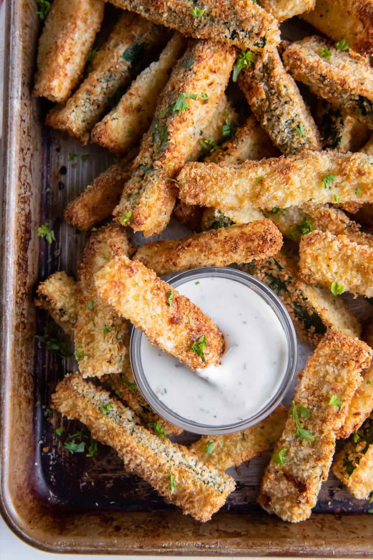 Air fryer zucchini fries on a baking sheet with one being dipped into the sauce.