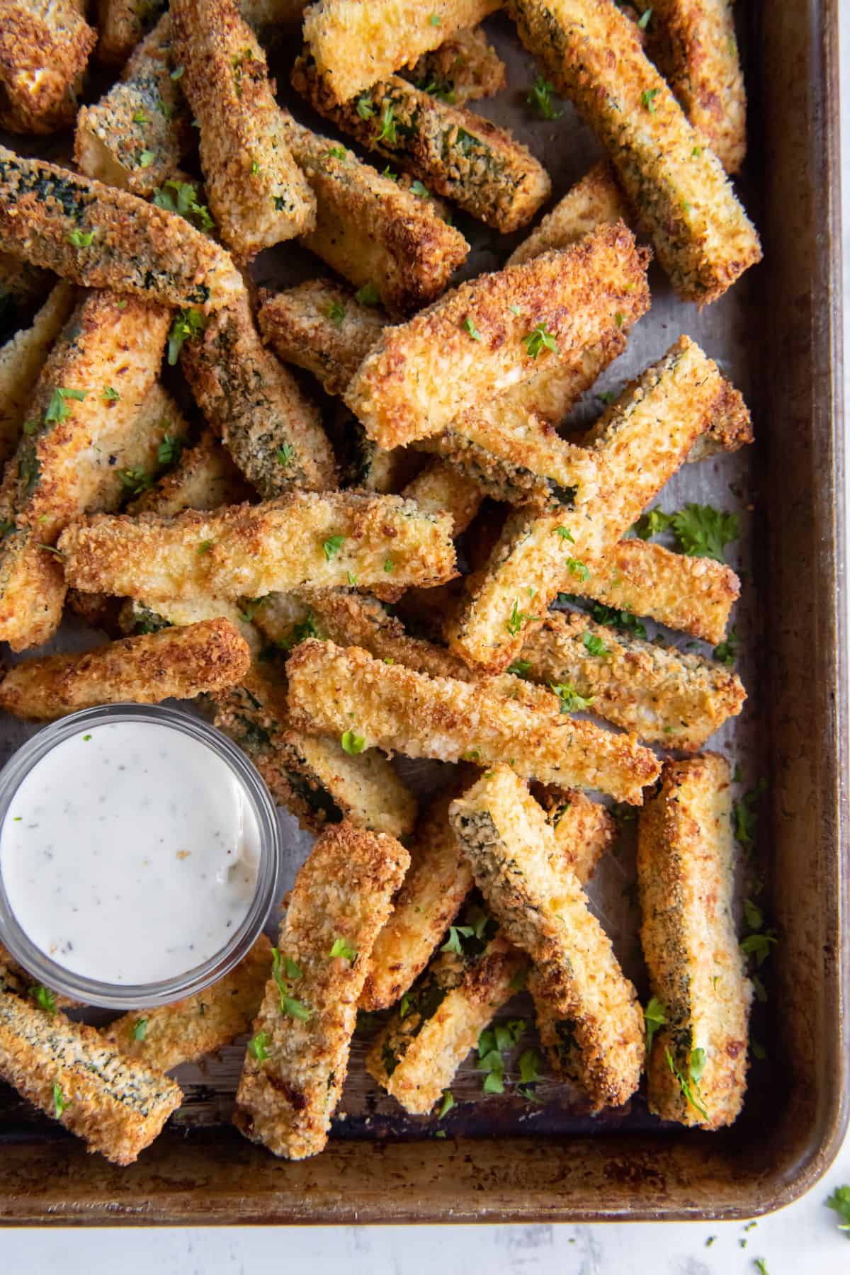 Overhead view of air fryer zucchini fries on a baking pan with a side of sauce.