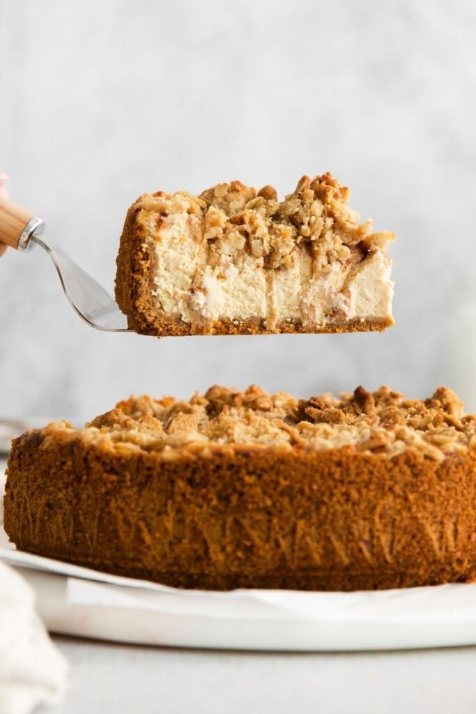 Apple crumble cheesecake from a side view with a slice of the cheesecake being held up by a cake spatula to see the layers.