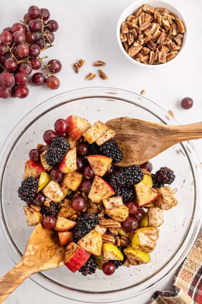 Tossing the fall fruit salad recipe in a clear glass bowl with wooden utensils.