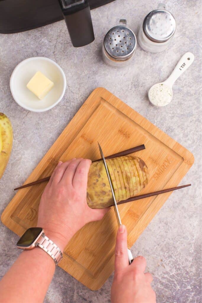 Potato on a cutting board with two chopsticks on the side cutting the potato into thin slices, but not all the way through.