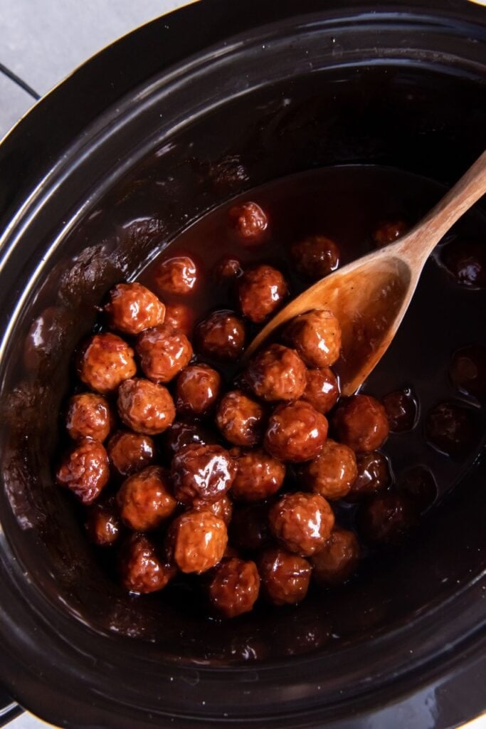 Cooked grape jelly BBQ meatballs in the slow cooker with a wooden spoon.