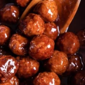 Close up of grape jelly BBQ meatballs in the crock pot with a wooden spoon.