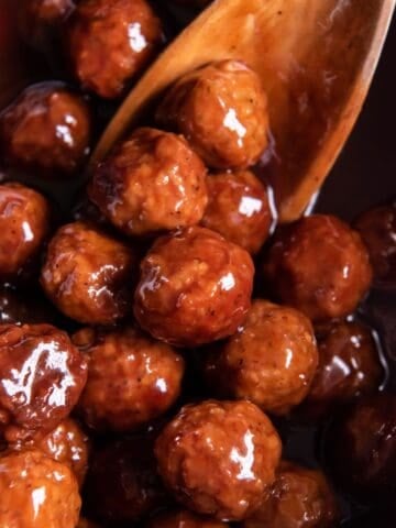 Close up of grape jelly BBQ meatballs in the crock pot with a wooden spoon.