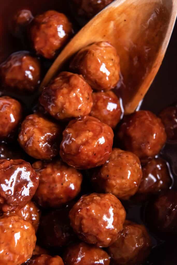 Close up of grape jelly BBQ meatballs in the crock pot with a wooden spoon.