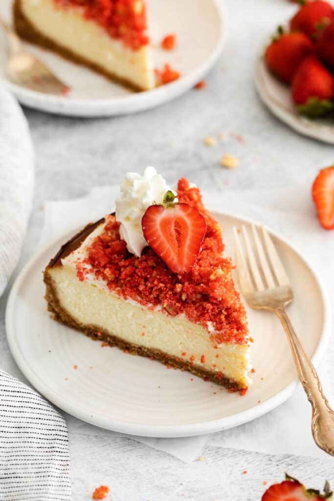 A slice of the strawberry crunch cheesecake recipe on a plate with a strawberry and whipped cream on it.