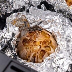 Close up of the roasted garlic in the air fryer basket with the foil partially unwrapped so you can see it fully roasted.