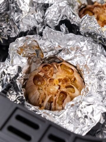 Close up of the roasted garlic in the air fryer basket with the foil partially unwrapped so you can see it fully roasted.