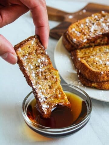 air fryer french toast sticks being dipped into a small bowl of maple syrup.