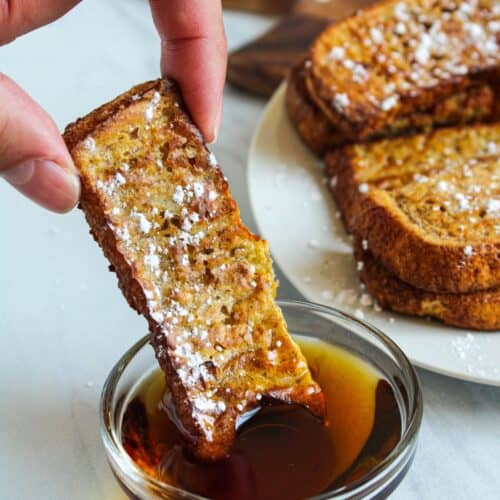 air fryer french toast sticks being dipped into a small bowl of maple syrup.