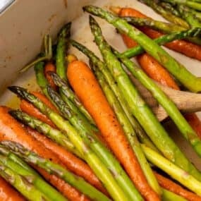 Roasting carrots and asparagus in a baking dish with a wooden spoon about to scoop some out.
