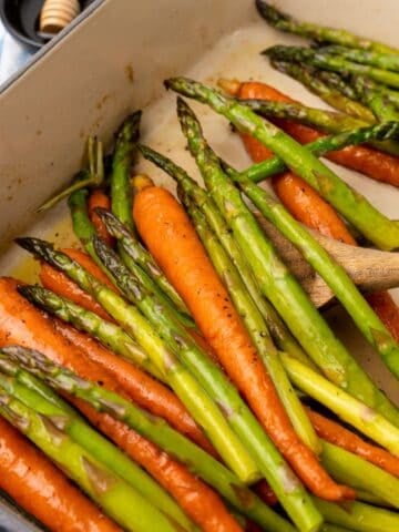 Roasting carrots and asparagus in a baking dish with a wooden spoon about to scoop some out.