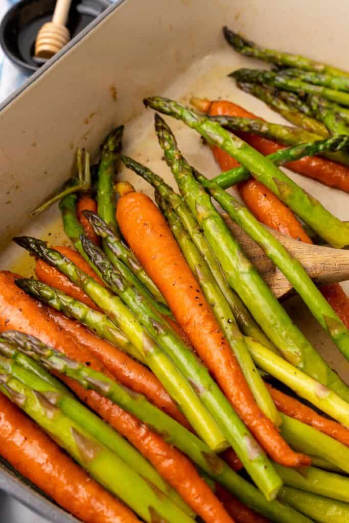 Roasting carrots and asparagus in a baking dish with a wooden spoon about to scoop some out.