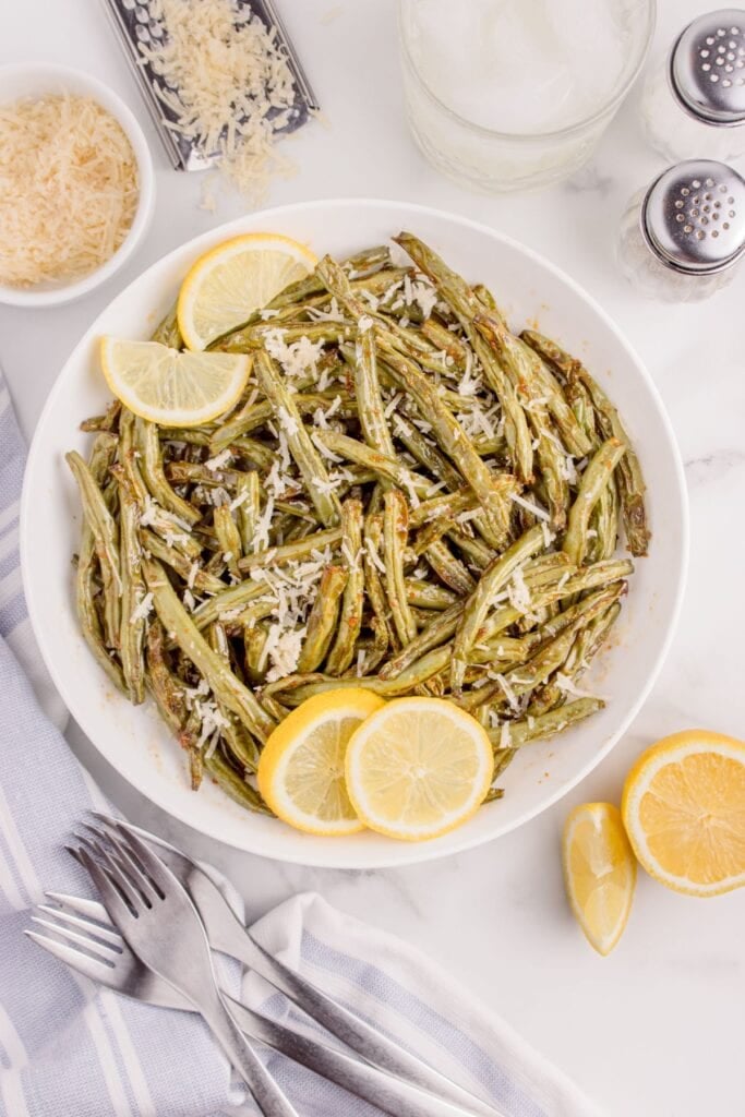 Cooked air fryer green beans topped with Parmesan cheese and lemon slices on the side.