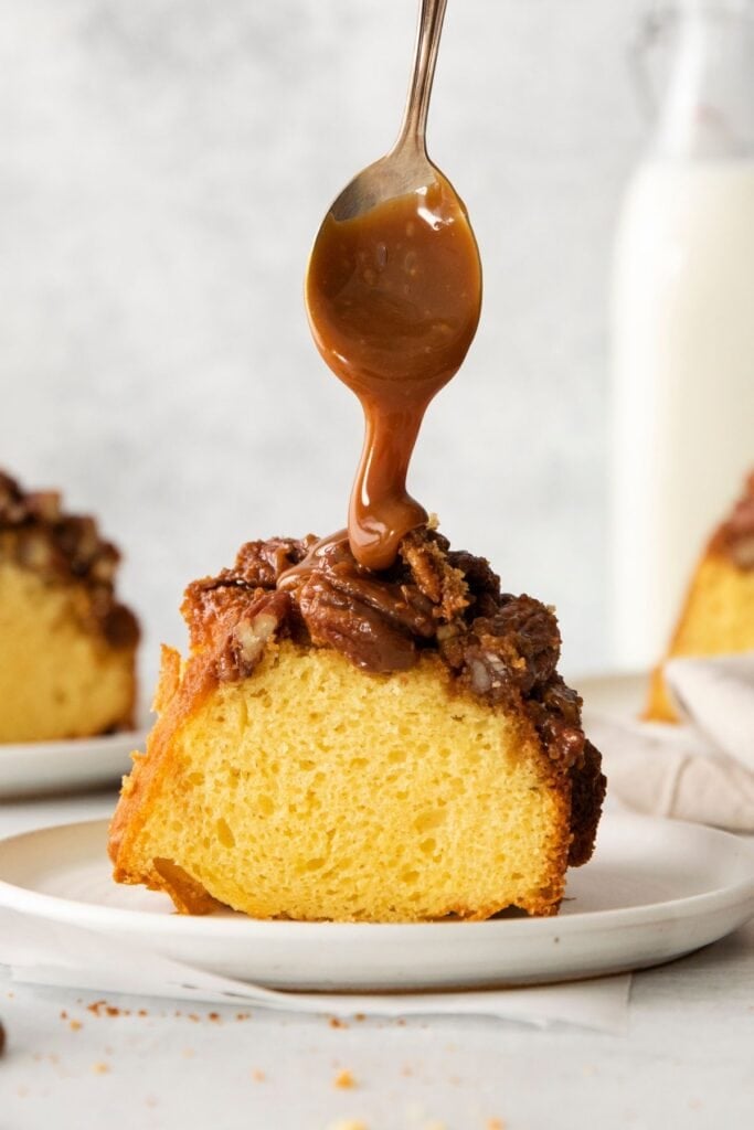 A slice of pecan pie cake on a plate with a spoon drizzling more caramel sauce on top.