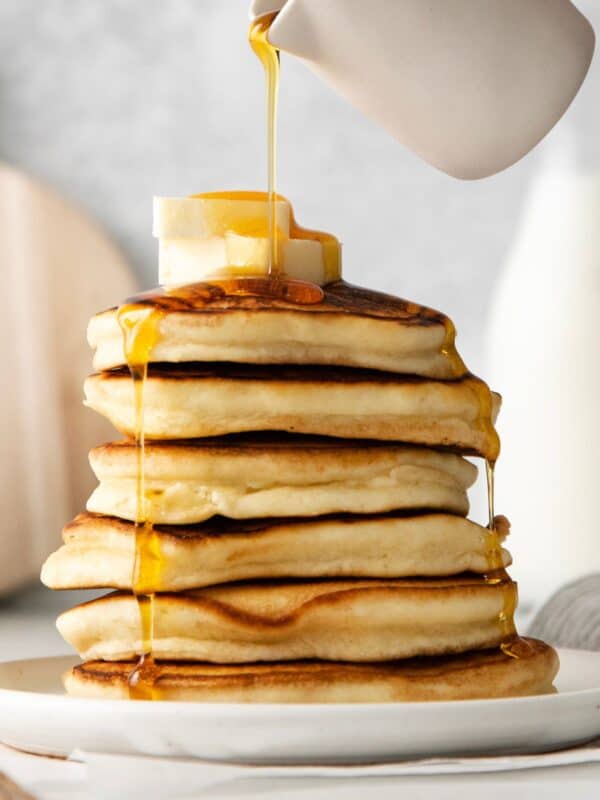 Pouring maple syrup over a stack of sweet cream pancakes.