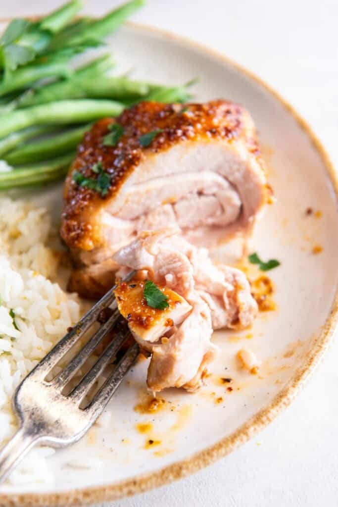 A fork holding a piece of boneless chicken thigh on a white plate with green beans in the background.