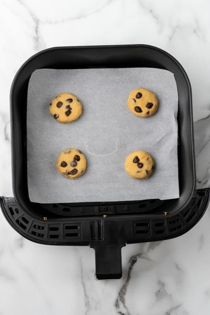 Overhead view of four scoops of cookie dough in a black air fryer basket lined with parchment paper.