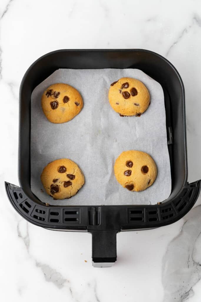 Overhead view of four cookies that have baked in an air fryer, resting on parchment paper in a black air fryer basket.