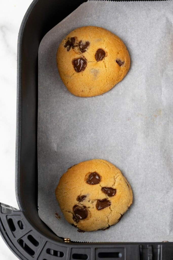 Close up view of 2 cookies on parchment paper resting in a black air fryer basket.