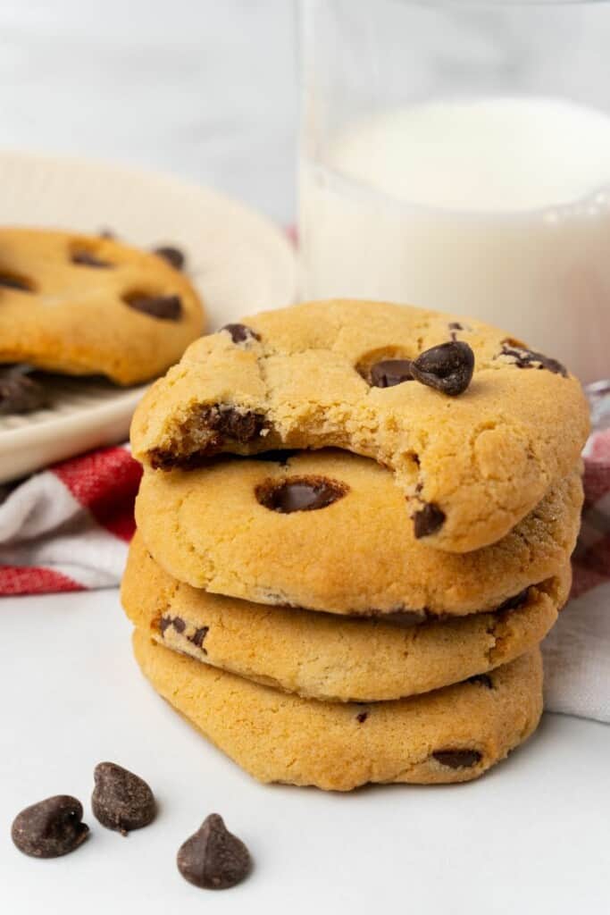 Stack of four air fryer chocolate chip cookies. The top cookie has a bite taken out, a glass of milk is in the background.