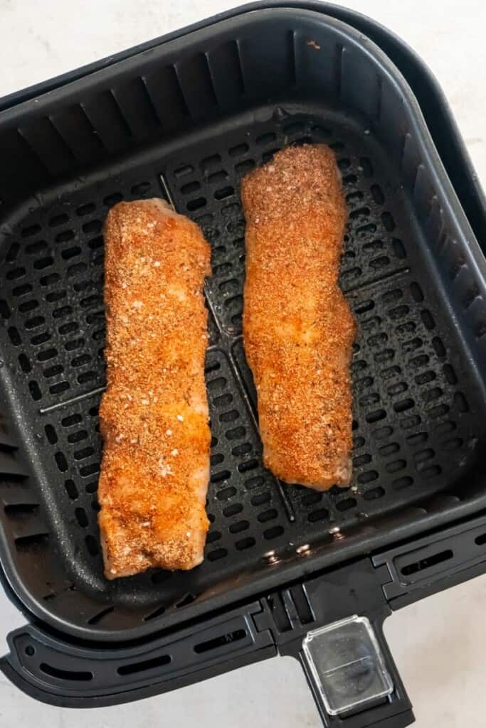 Frozen Cod with spices rubbed on in a black air fryer basket.