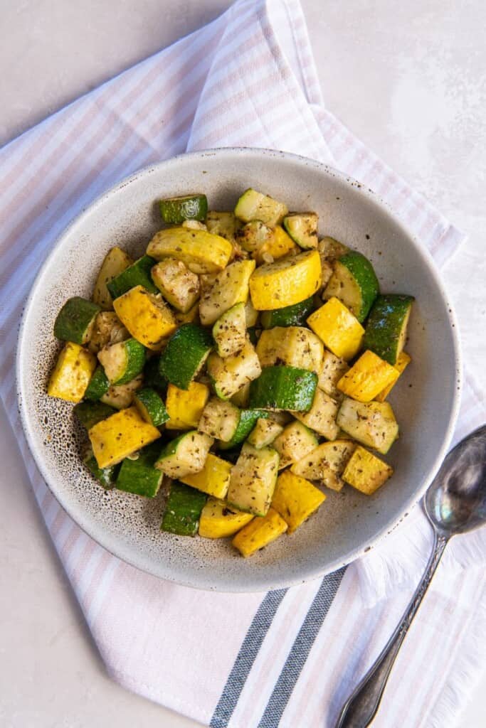 Zoomed out overhead view of squash and zucchini in a white bowl with a spoon.