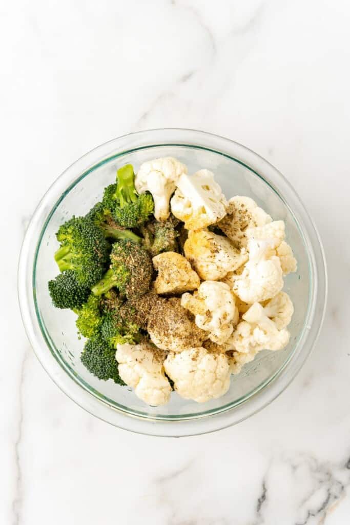 Broccoli and Cauliflower in a clear mixing bowl topped with with olive oil, Italian seasoning, garlic powder and salt.