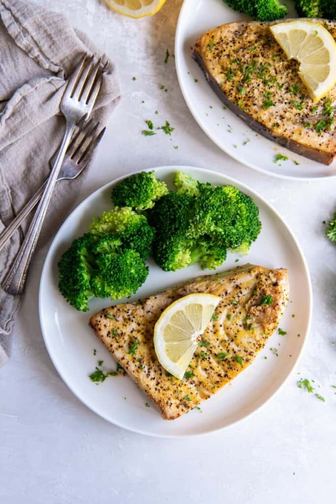 Prepared air fryer swordfish served on a white plate topped with a lemon wedge, next to broccoli.
