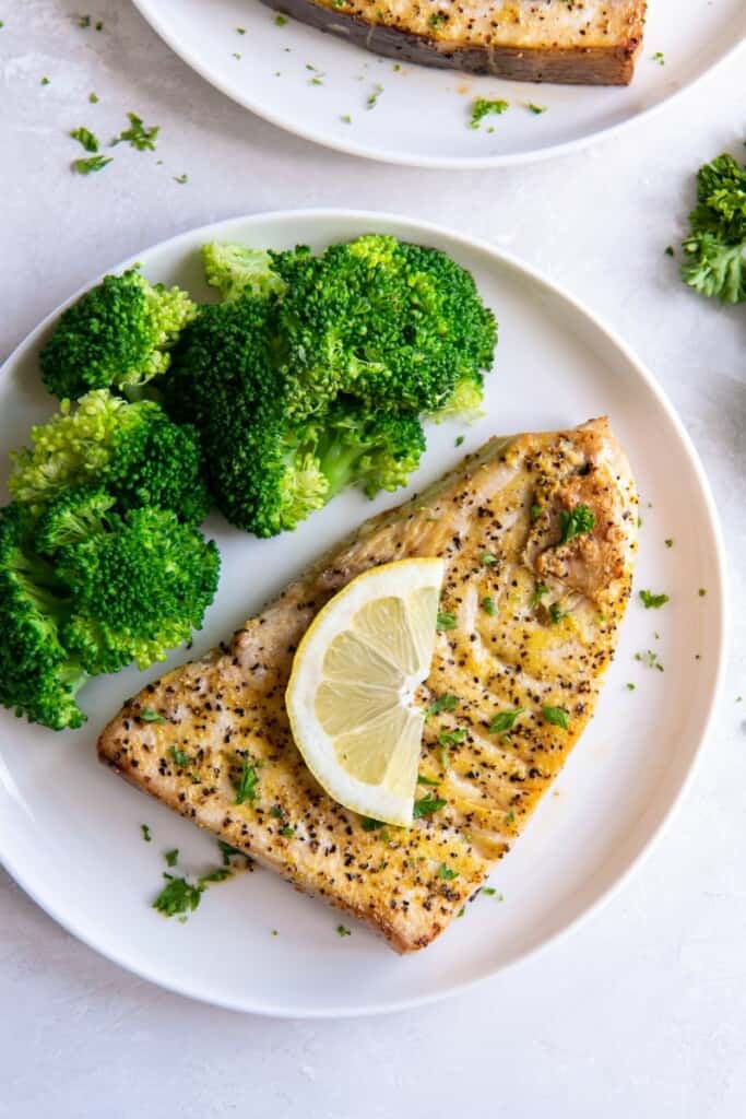 Closeup of prepared air fryer swordfish steak topped with a lemon wedge on a white plate with broccoli.